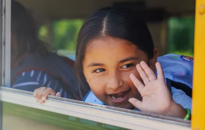 Young female student waving her hand outside a bus window.