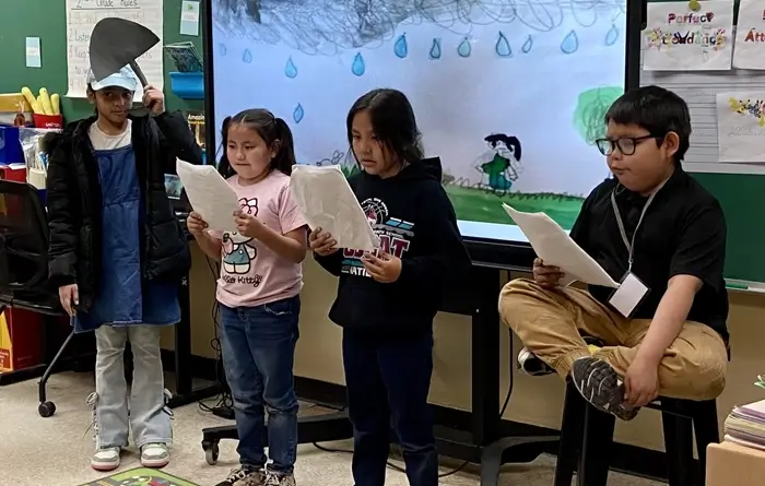 Children perform a classroom skit while reading from papers in front of a rainy scene on a smartboard.