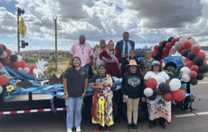 A group of people pose in front of a colorful parade float decorated with red, white, black, and blue balloons, with a cloudy sky and rural landscape in the background.