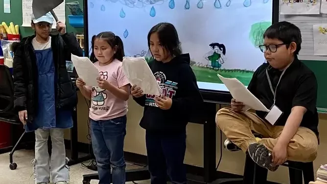 Children perform a classroom skit while reading from papers in front of a rainy scene on a smartboard.