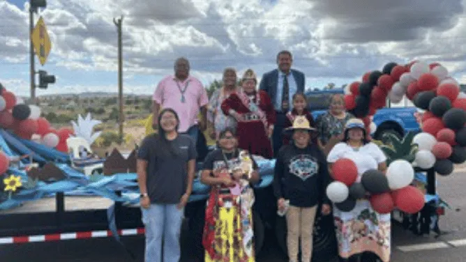 A group of people pose in front of a colorful parade float decorated with red, white, black, and blue balloons, with a cloudy sky and rural landscape in the background.